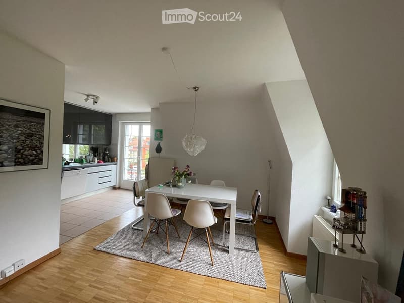 Bright, open-plan living and dining area with wooden flooring, white dining table, modern chairs, kitchen area with cabinets and appliances, and decorative items including a framed picture and a vase with flowers.