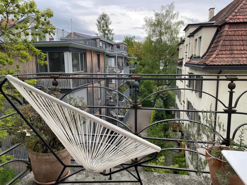 Balcony with a white hanging chair, small table, and potted plants, overlooking a residential area with modern and traditional buildings.