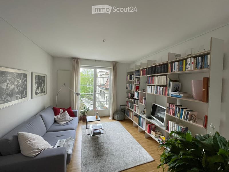 Modern living room with a grey sectional sofa, coffee tables, a large bookshelf filled with books, a television, a floor lamp, and decorative plants.