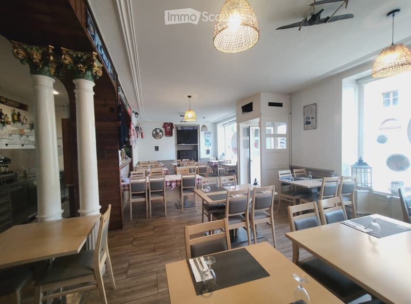 Restaurant interior with wooden tables, chairs, and place settings, featuring hanging lamps and large windows.