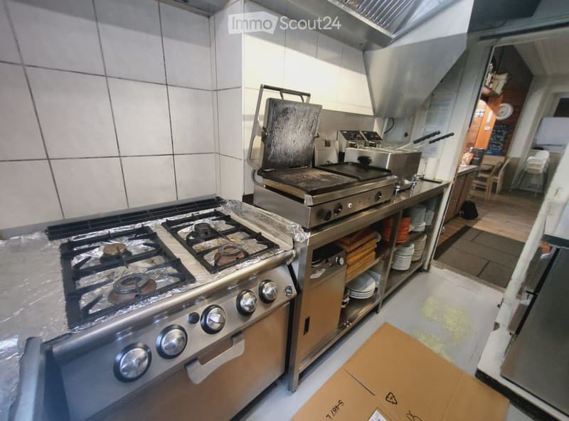 Commercial kitchen with stainless steel appliances including a gas stove, griddle, and oven. Shelves hold plates and utensils, with a cardboard box on the floor.