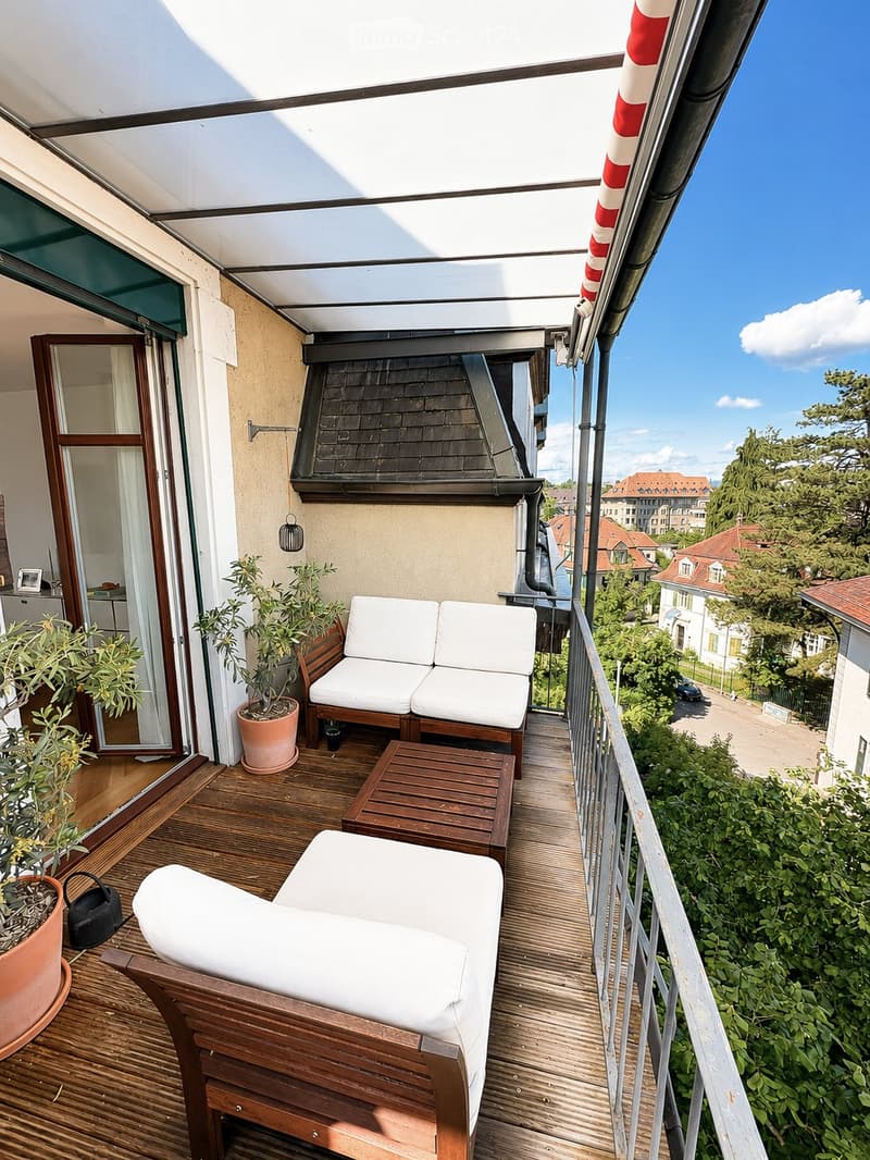 Balcony with wooden decking, white sofa, wooden table, lounge chair, and potted plants, overlooking a cityscape with trees and buildings.