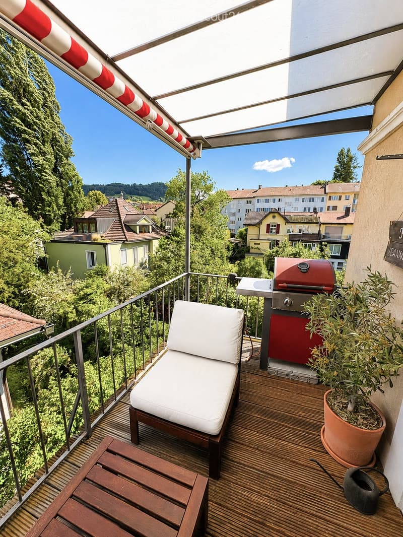 Balcony with wooden decking, white lounge chair, small table, red grill, potted plant, and a view of the neighborhood.