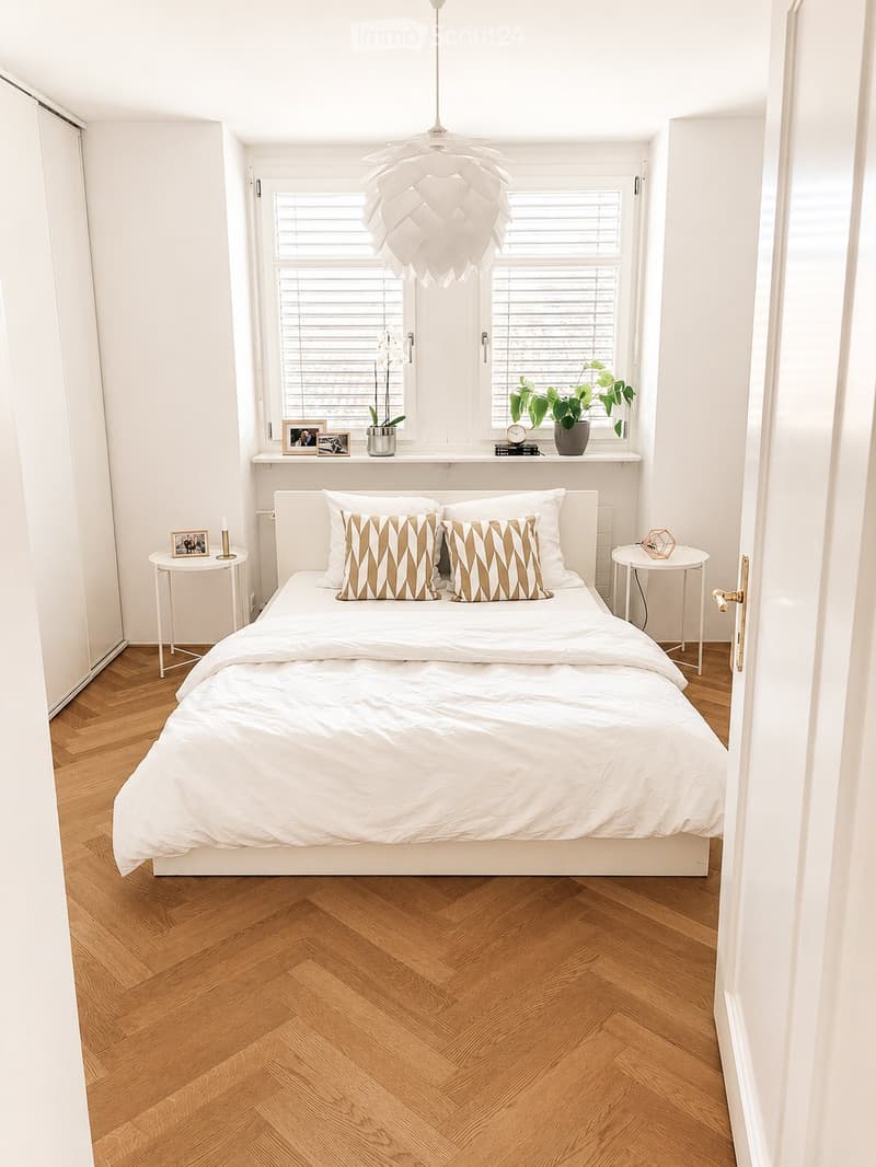 Modern bedroom with a white bed, patterned pillows, wooden herringbone flooring, and a large window with blinds. Side tables with lamps and potted plants are visible.