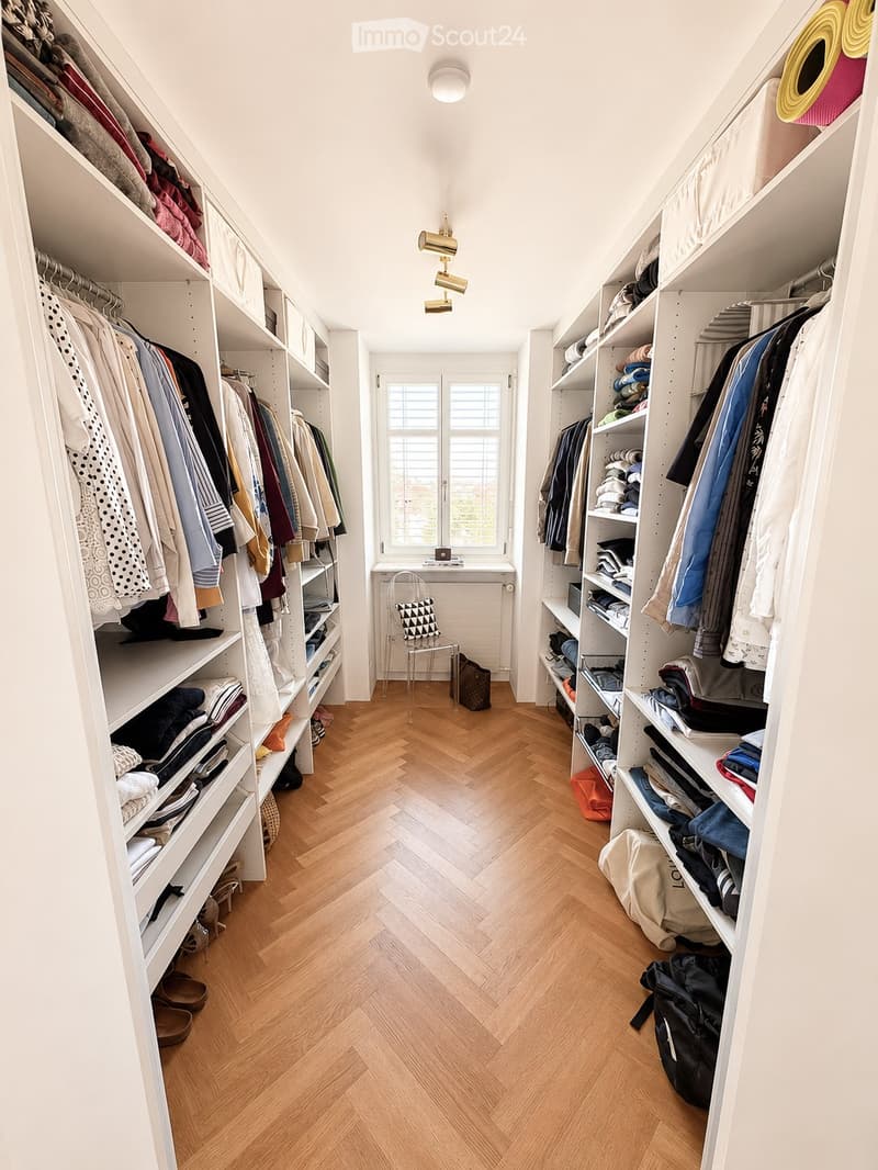 Spacious walk-in closet with built-in white shelving units, wooden herringbone flooring, and large window with blinds. Contains neatly organized clothing, shoes, and accessories.