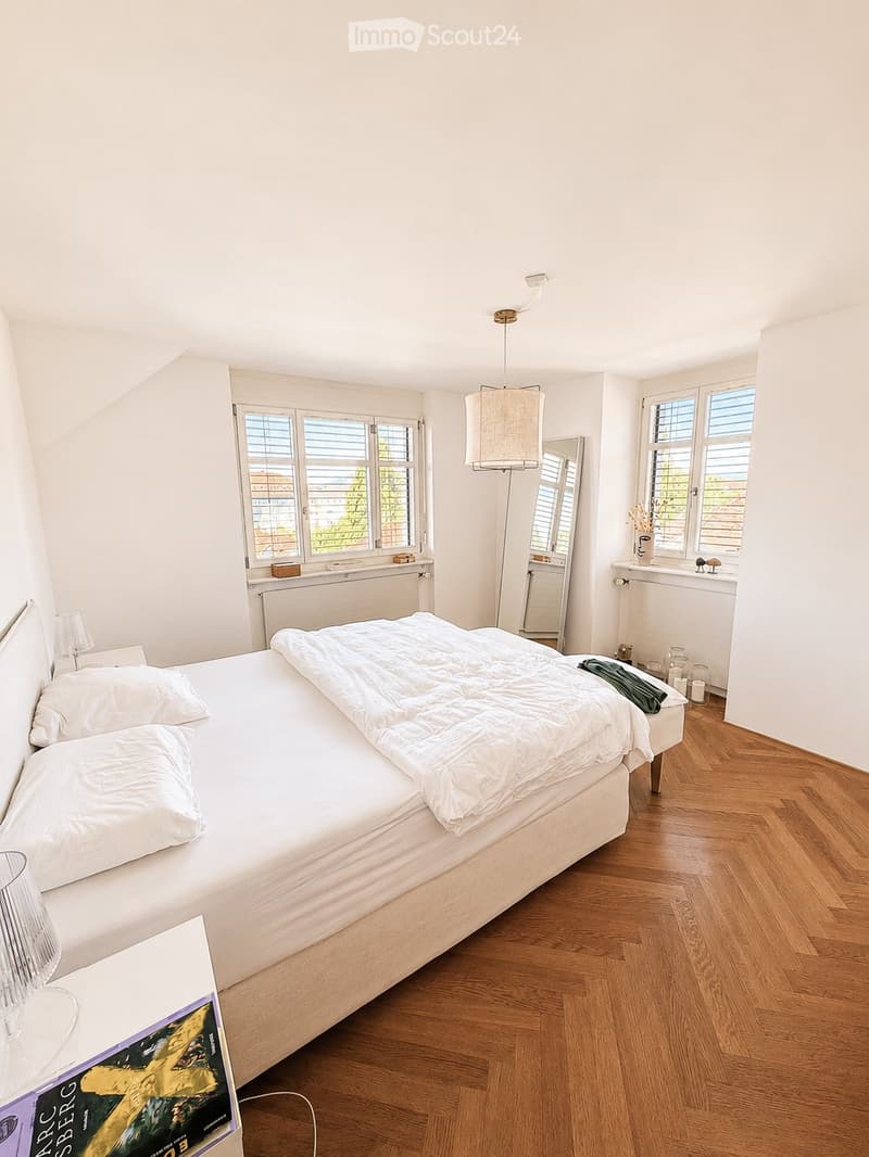 Bright bedroom with wooden flooring, large bed with white bedding, mirror, table lamp, and decorative items on windowsills.