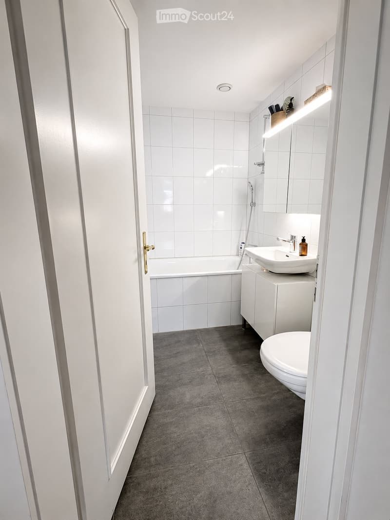 Modern bathroom with white tiles, a bathtub, sink, toilet, and a mirrored cabinet. The floor is tiled in a dark gray color, and the walls are white.