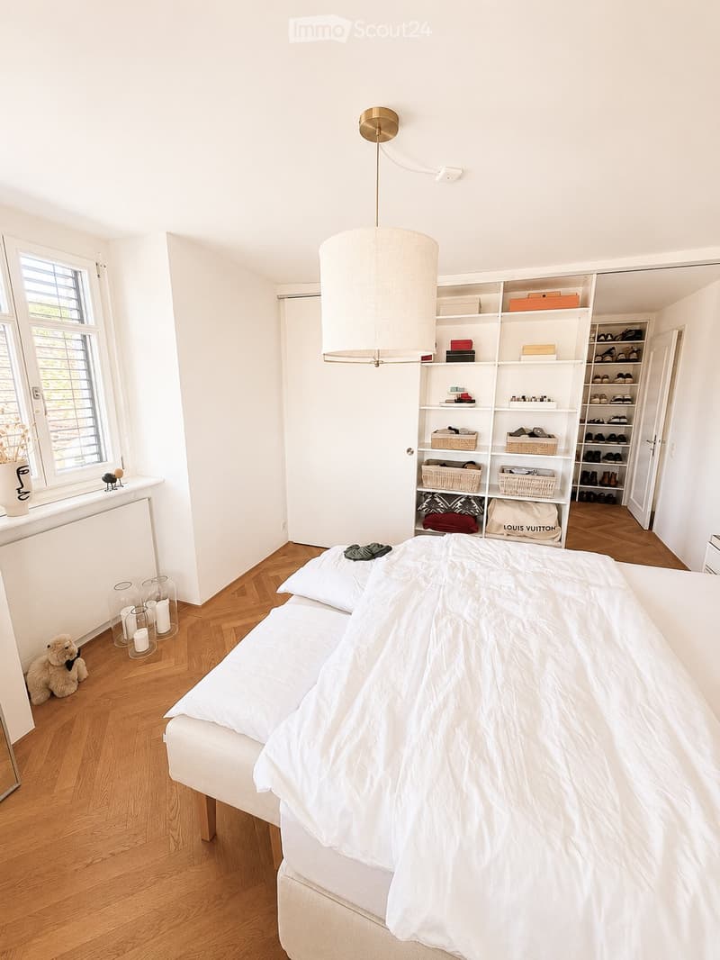 Bright bedroom with a white bed, wooden floor, built-in shelves, and decorative candles near the window.