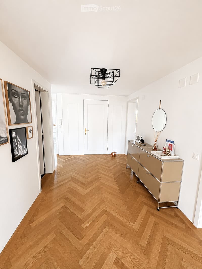 Hallway with herringbone wooden flooring, white walls, modern lighting fixture, dresser with mirror, and artwork on the wall.