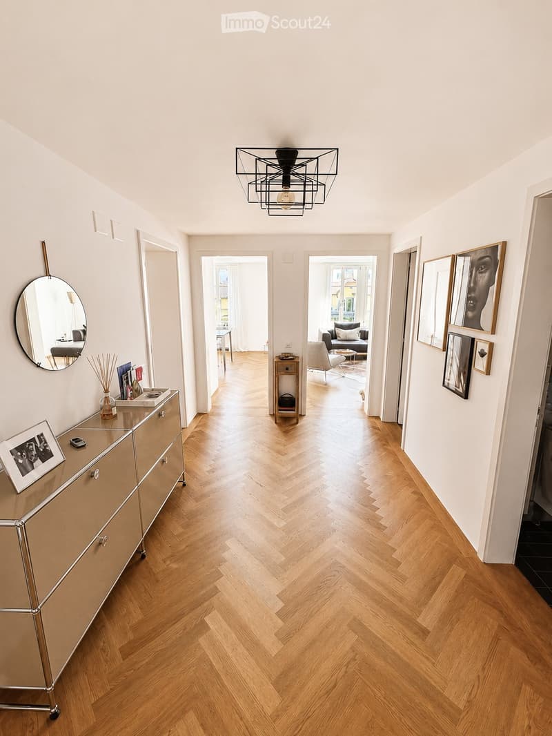 Elegant hallway with herringbone wooden flooring, modern lighting fixture, and framed artwork on the walls. A small console table with decorative items is visible on the left, and a mirror is mounted on the wall.