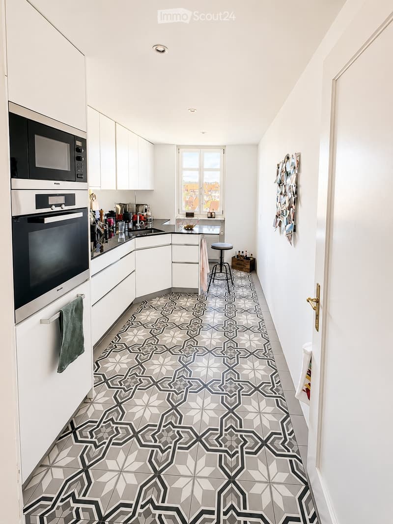 Modern kitchen with white cabinets, stainless steel appliances, and a geometric patterned tile floor. Features a microwave, oven, sink, and a small stool near the window.