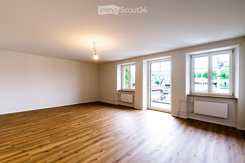 Empty room with wooden flooring, white walls, and multiple windows allowing natural light. Radiators are visible along the walls.