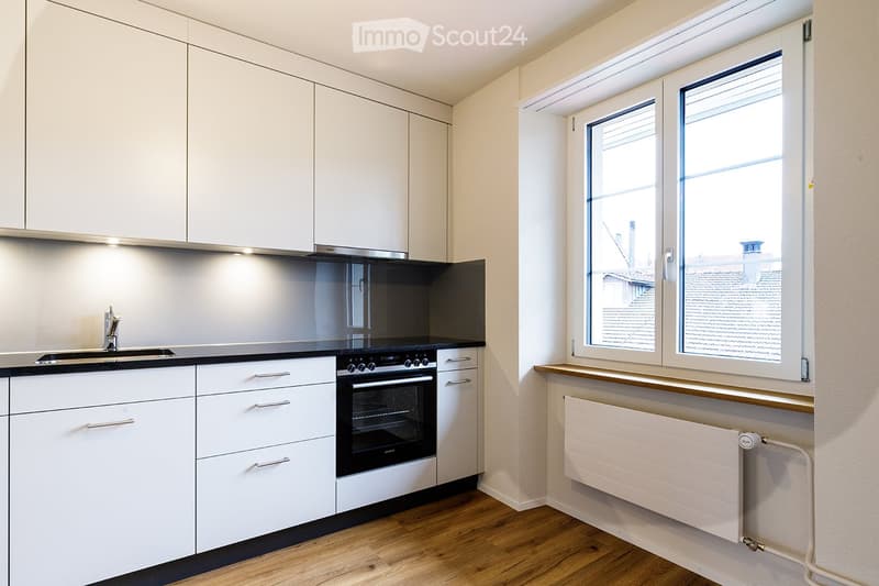 Modern kitchen with white cabinets, stainless steel appliances, and a black countertop. Features a sink with a modern faucet, an oven, and a radiator under the window.