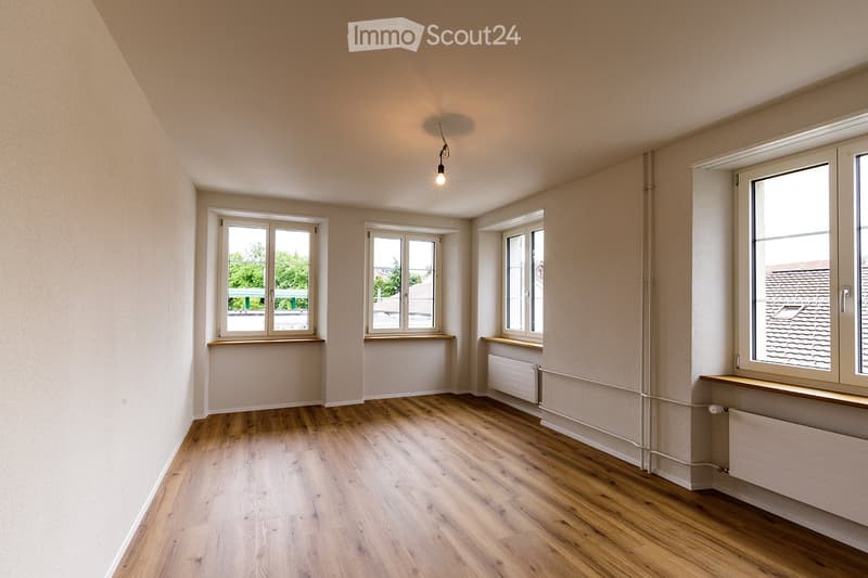 Empty room with wooden flooring, white walls, multiple windows, and radiators. No furniture or equipment present.