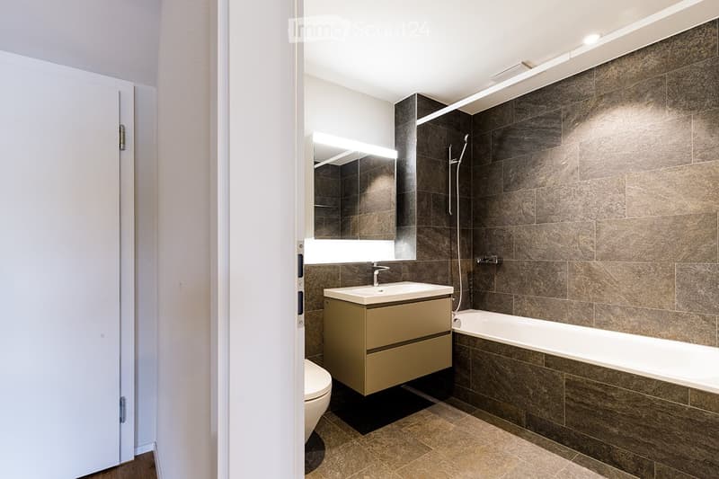 Modern bathroom with a beige vanity, large mirror, bathtub, and toilet. Dark brown tiles on walls and floor.