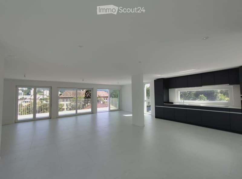 Open-plan unfurnished space with white tiled flooring, large sliding glass doors leading to a balcony, and a modern kitchen area with black cabinetry and a white countertop.