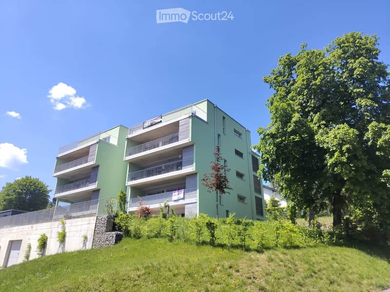 Modern multi-story apartment building with green and white exterior, multiple balconies, surrounded by trees and greenery.