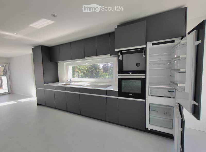 Modern kitchen with dark grey cabinets, white countertops, stainless steel appliances, and a large window overlooking greenery.