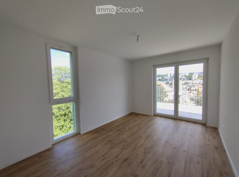 Empty room with wooden flooring, white walls, large windows, and sliding glass doors leading to a balcony.