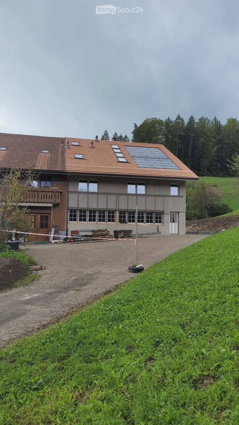 Two-story house with a red-tiled roof, large windows, and a balcony on the upper level. The house is surrounded by a grassy area and trees.