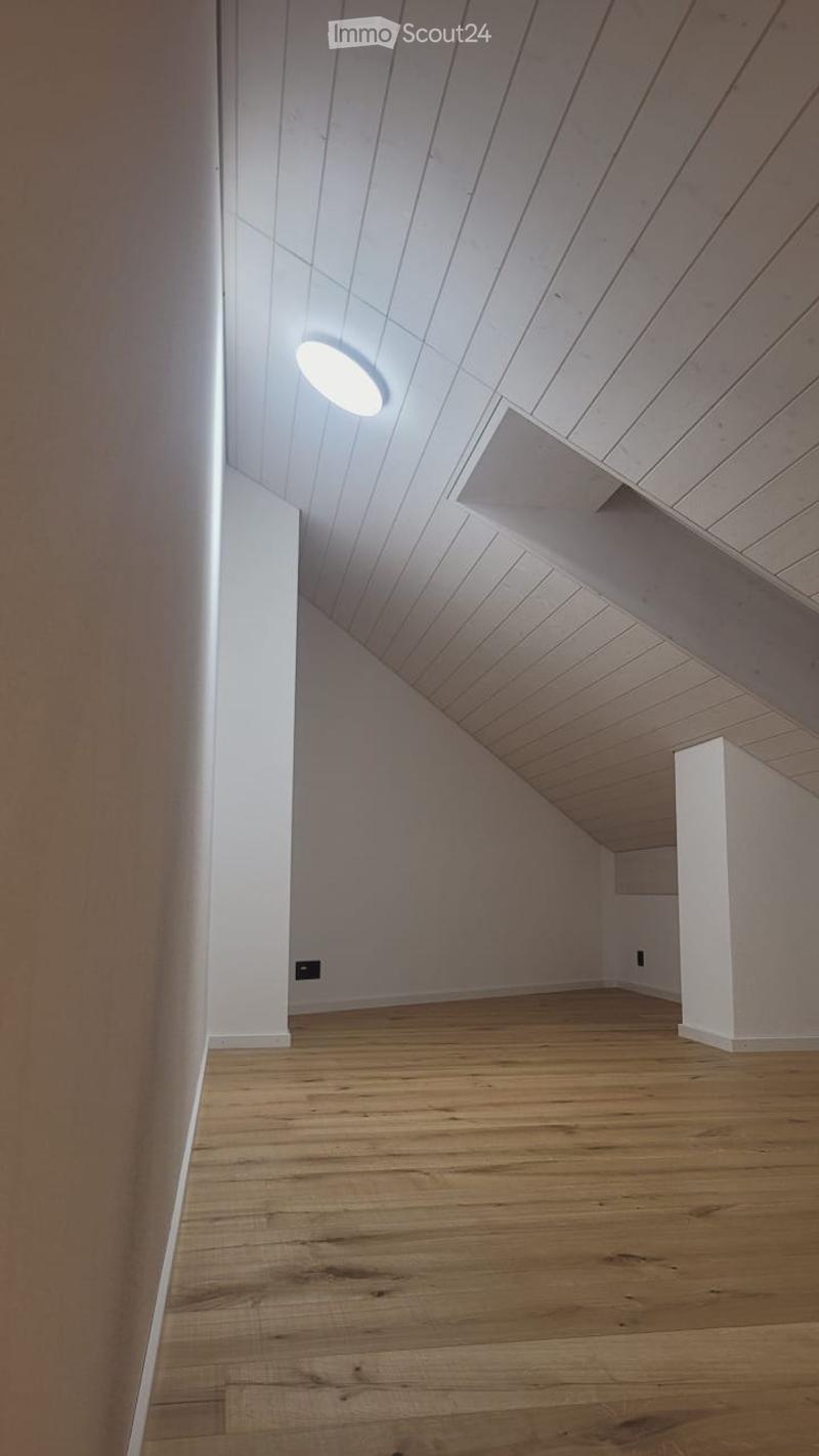 Empty room with wooden flooring, white walls, and a slanted wooden ceiling with a single round light fixture.