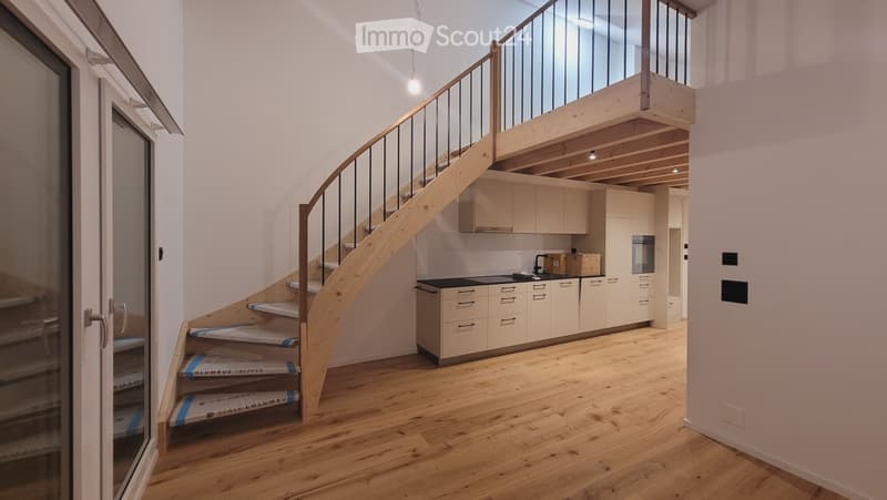 Modern hallway with wooden flooring, a curved staircase leading to an upper level, and a small kitchen area visible in the background.