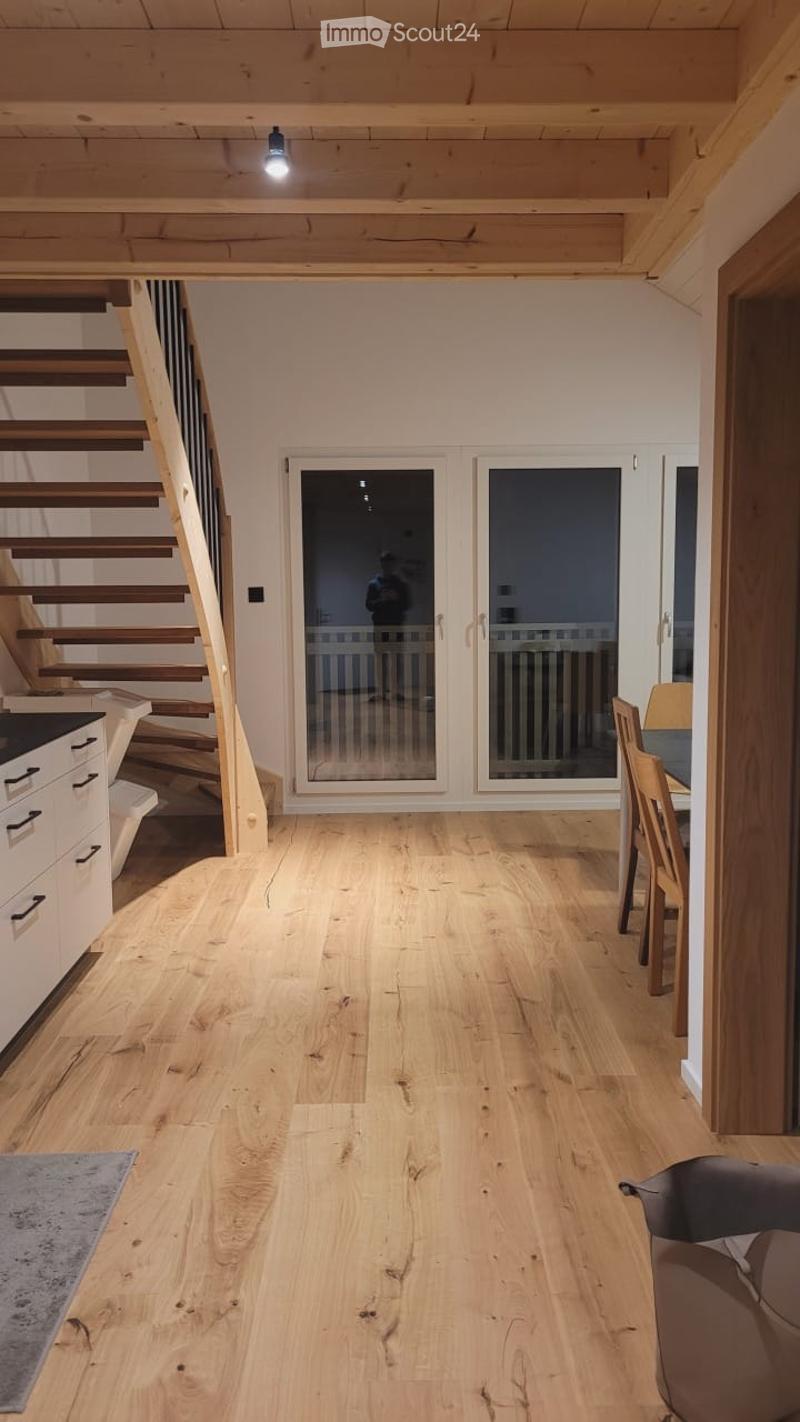 Bright, open-plan living and dining area with wooden flooring, exposed wooden beams, and large glass doors leading outside. Includes a sofa, dining table with chairs, and a kitchen cabinet.