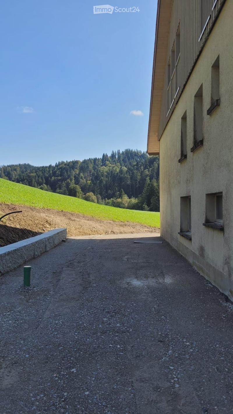 Gravel driveway leading to a modern building with a raised ground floor, surrounded by green hills and trees.