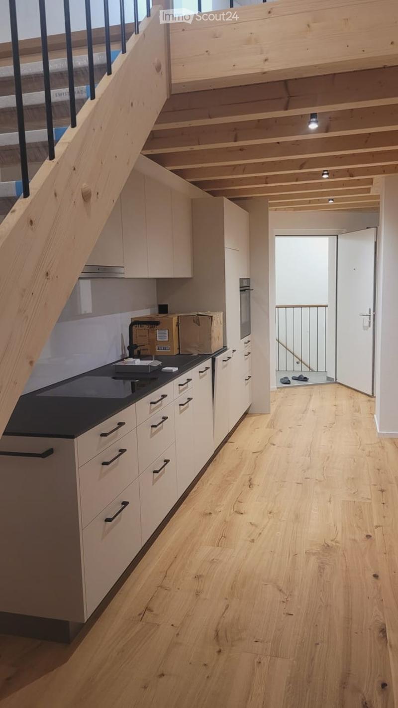 Modern hallway with wooden flooring, white walls, and a staircase leading to an upper level. A kitchen area is visible to the left with white cabinets, a black countertop, and a built-in oven. A small balcony or terrace is accessible through an open doorw