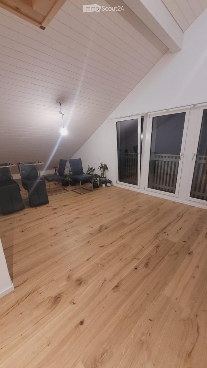 Bright attic room with wooden flooring, slanted white ceiling, large sliding glass doors leading to a balcony, and a few modern chairs and a small table with potted plants.