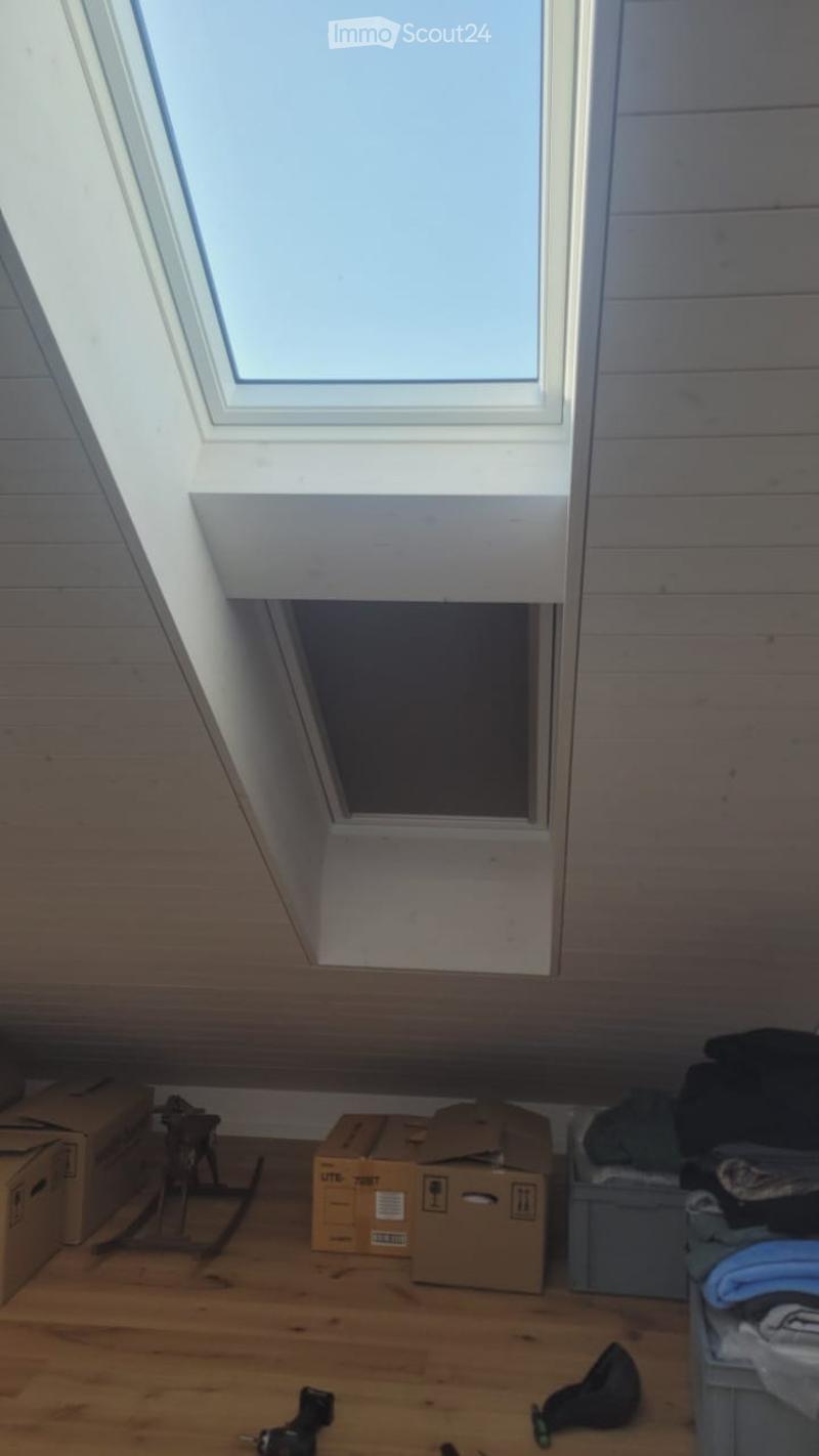 An attic space with wooden walls and floor, featuring a skylight window and several cardboard boxes and items scattered on the floor.