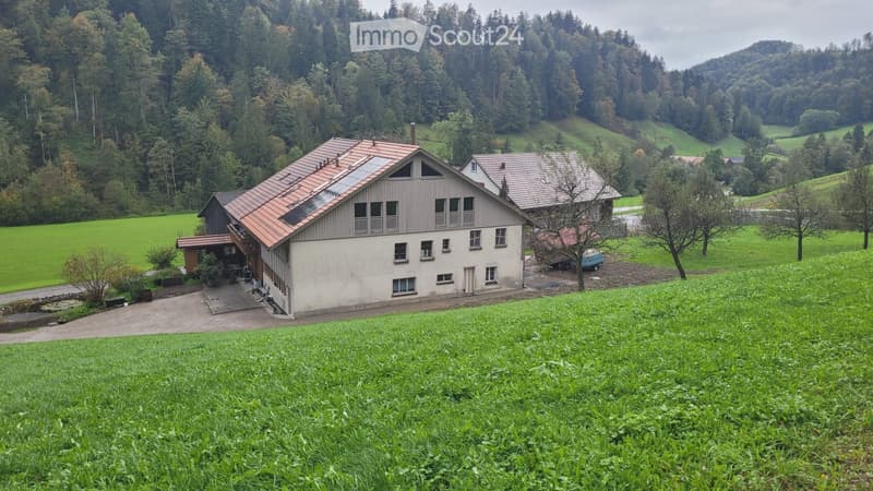 A two-story house with a red-tiled roof and solar panels, surrounded by green fields and trees in a rural setting.