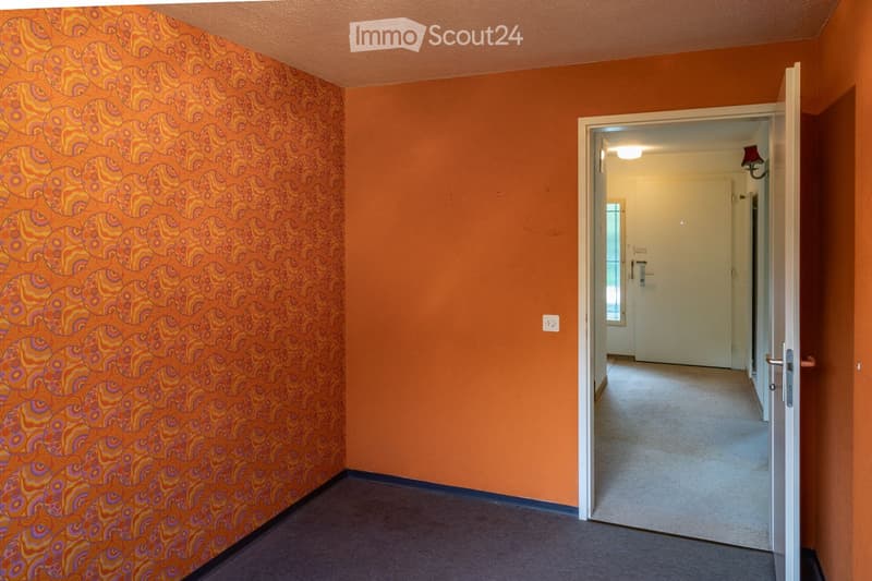 A narrow hallway with orange walls and patterned wallpaper on one side, leading to an open doorway that reveals another room with white walls and a light fixture.
