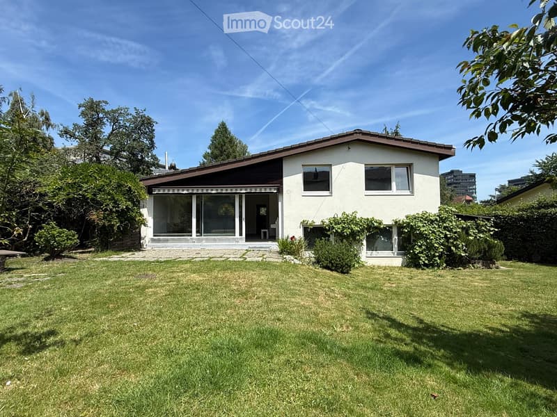 Single-story house with white walls, dark roof, large windows, and a covered entrance area. The house is surrounded by a well-maintained lawn and trees.
