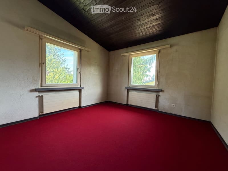 Empty room with red carpet, two windows, radiators, and wooden ceiling beams.