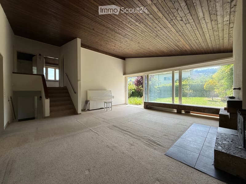 Bright, open living space with a high, slanted wooden ceiling, large windows, a staircase leading to an upper level, and a white piano near the window.