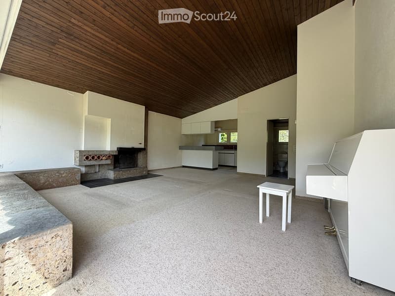 Open-plan living space with a fireplace, a white piano, a small white stool, and a kitchen area visible in the background.