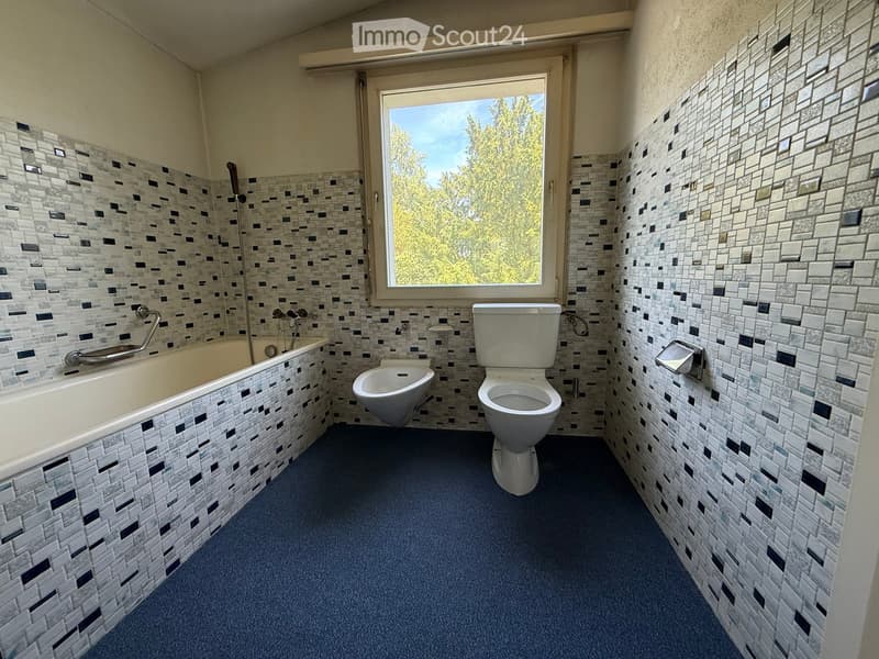 Bathroom with a bathtub, toilet, bidet, and a large window overlooking trees. The walls are tiled with a mosaic pattern, and the floor is carpeted.