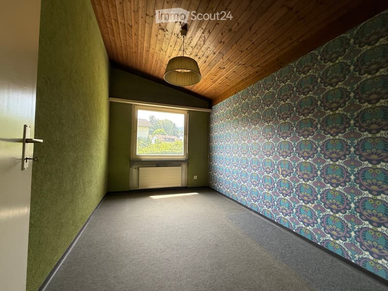 Empty room with wooden slanted ceiling, grey carpet, green walls, and a large window with view of houses and trees.