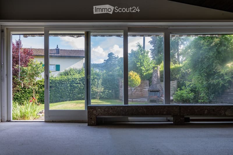 Empty room with large glass windows overlooking a garden, a stone bench placed in front of the windows, and a view of a brick chimney and greenery.