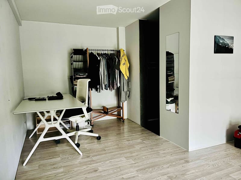 A modern, minimalist room with a white desk, a white chair, a clothing rack with hanging clothes, a mirror, and a picture frame on the wall.
