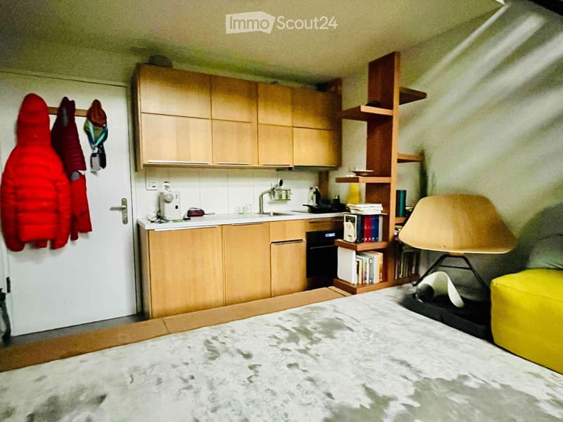 A furnished room with a bed, a chair, a yellow stool, a wooden cabinet, a shelf with books, and a small kitchen area with a sink, faucet, and oven.