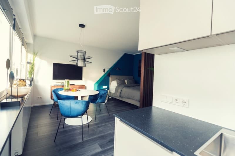Modern, open-plan living space with a kitchen area, dining table, and a separate bedroom area. Features include a sleek black countertop, a white dining table with four blue chairs, a television, a bed with pillows and a duvet, and a stylish hanging lamp.