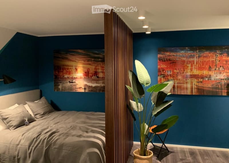 Modern bedroom with blue walls, a large bed with pillows, a potted plant, a chair, and two large wall art pieces.
