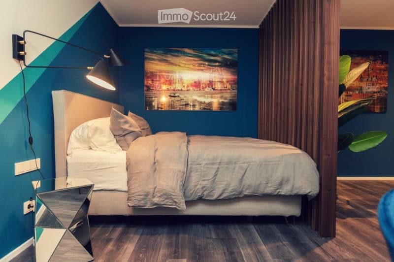 Modern bedroom with a large bed, nightstand, mirror, wall art, and a stylish lamp. The room features dark blue walls and wooden flooring.