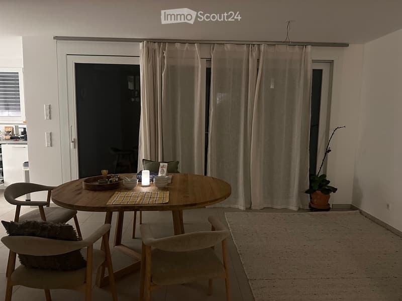 A modern dining area with a round wooden table, four chairs, a lamp, and a potted plant near the window.