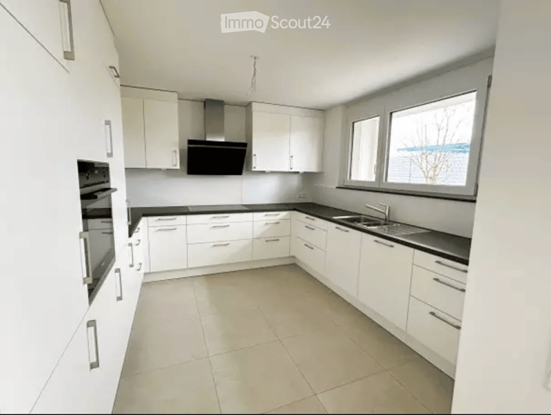 Modern kitchen with white cabinets, black countertops, and stainless steel appliances including an oven and range hood. Large windows provide natural light.