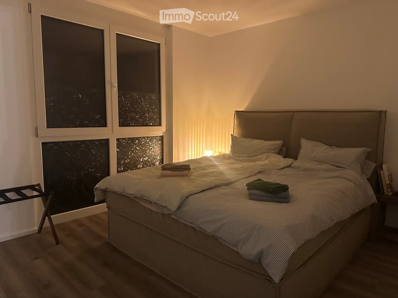 Modern bedroom with a large bed, nightstand, and a small chair near the window. The room features large windows with a view and warm lighting.