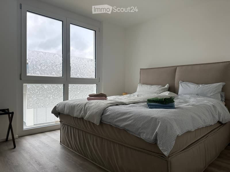 Modern bedroom with large windows, a neatly made bed with pillows, a headboard, and folded towels on the bed.