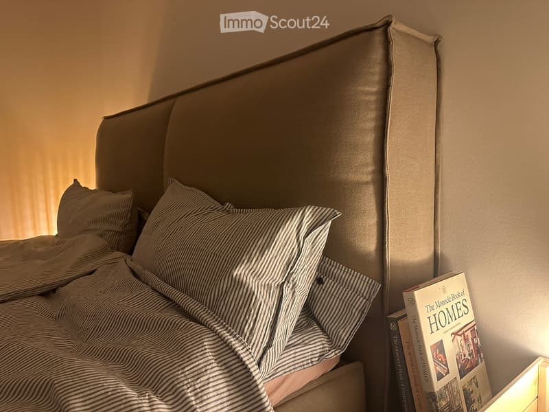 A neatly made bed with a headboard, pillows, and a striped blanket, accompanied by a stack of books on a bedside table.